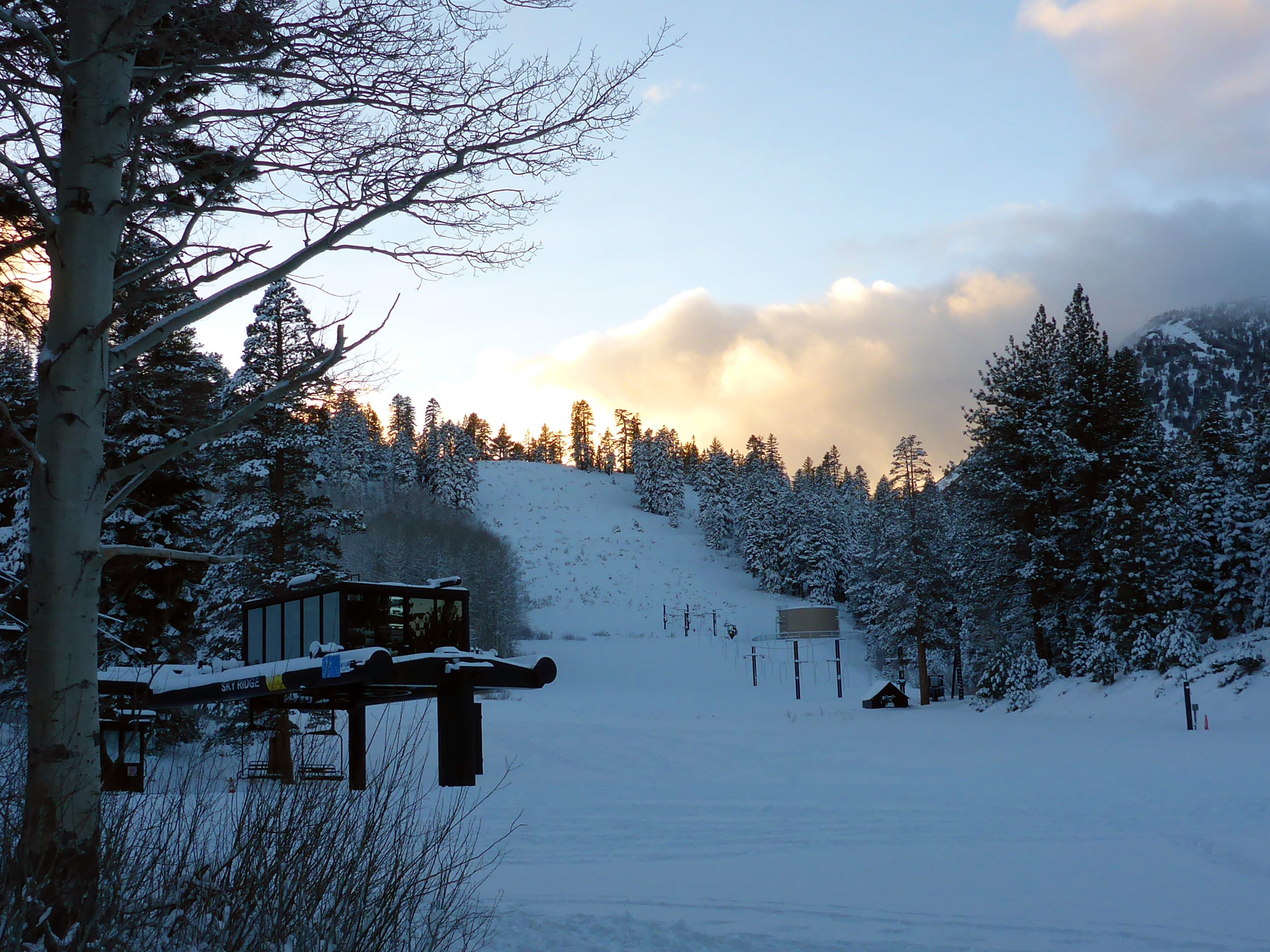 Snow-covered base area with a chairlift terminal and a cut ski run rising into pines at dusk