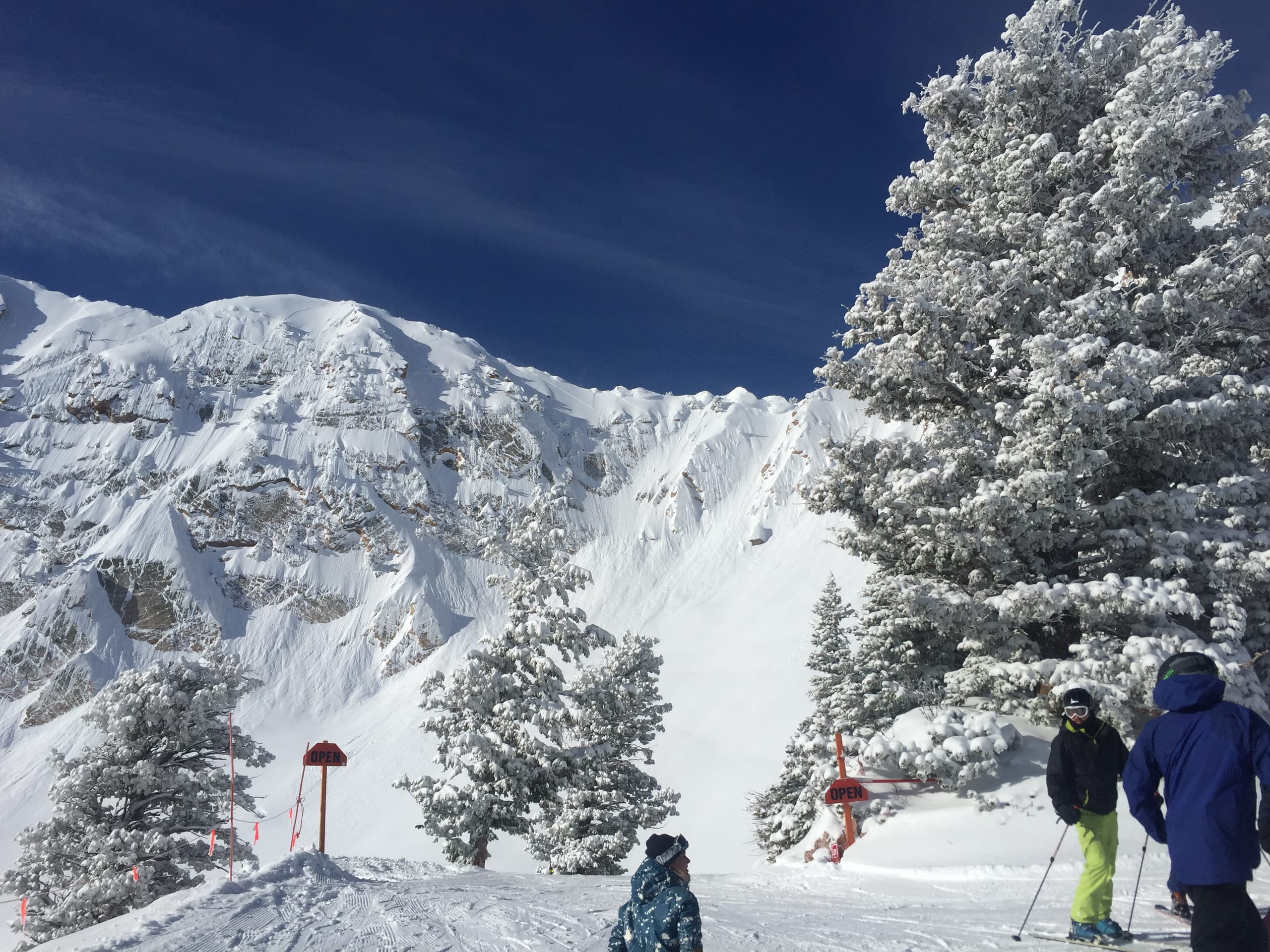 Skiers on a groomed run below a snow-covered cirque and rime-coated pines on a sunny day