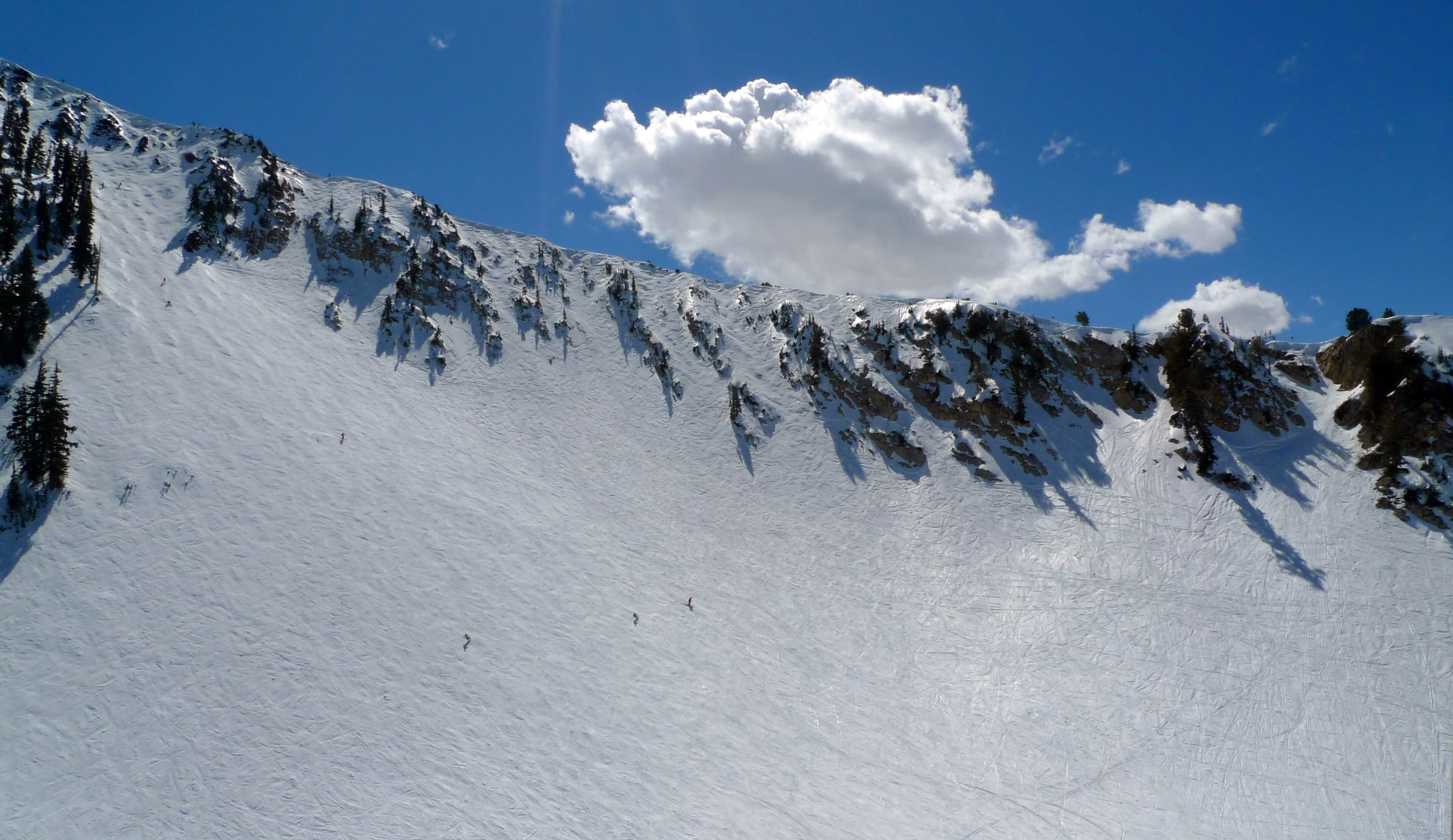 Snow-covered cirque bowl with rocky ridgeline and tiny skiers on the wide open slope under blue sky