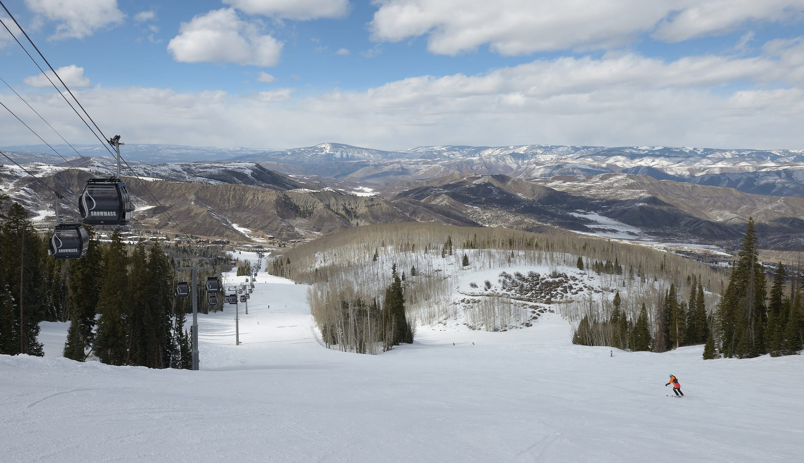 Groomed run at Snowmass with gondola cabins overhead, a lone skier, and distant snowy peaks