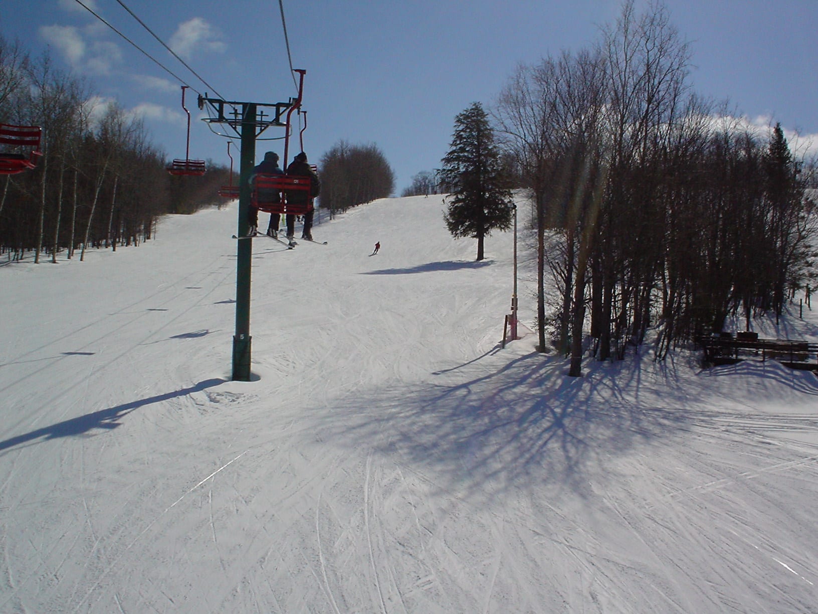 Skiers ride a chairlift above a snow-covered run lined with bare trees at a Michigan ski area