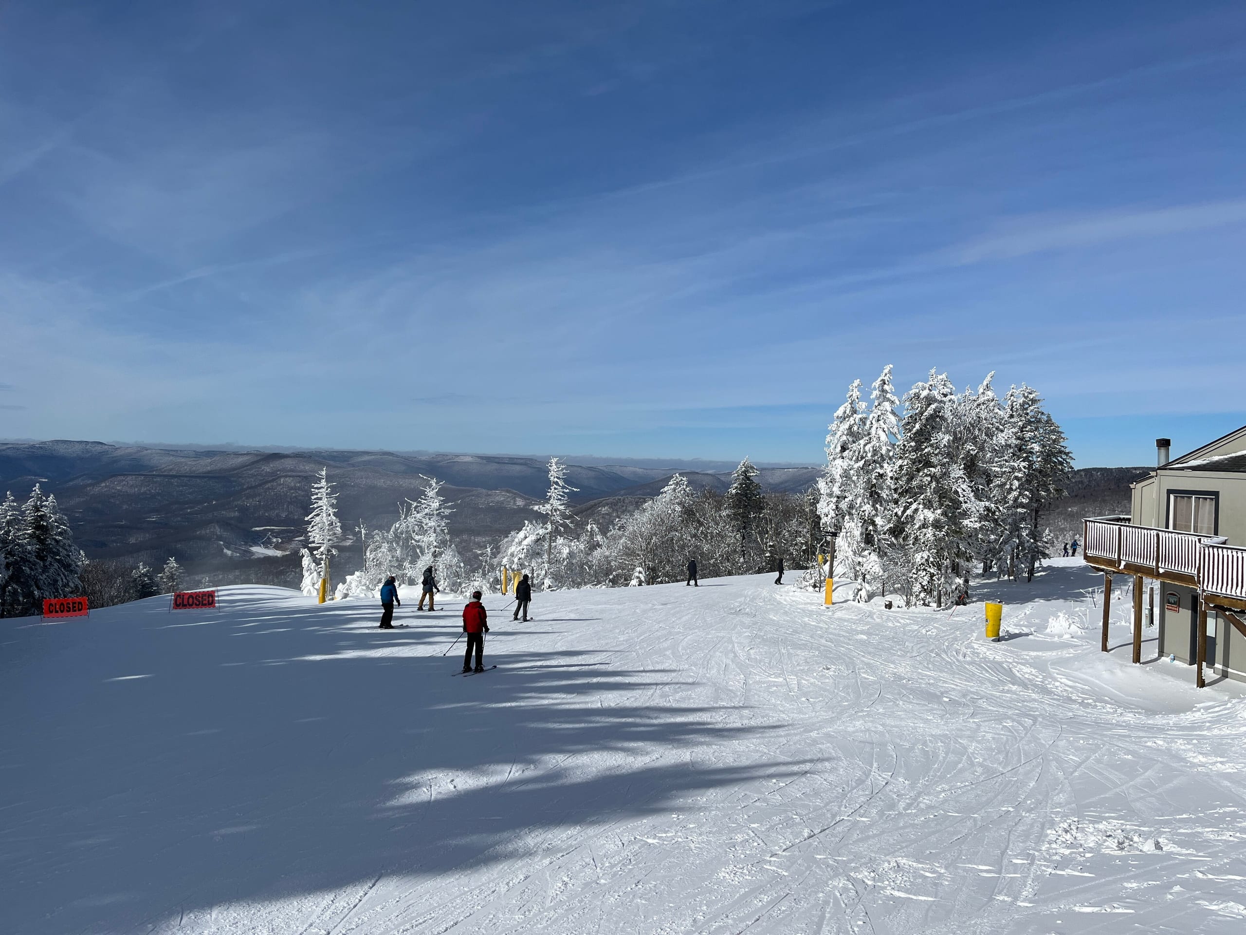 Skiers at a snow-covered summit trailhead with rime-coated trees and distant ridges under blue sky