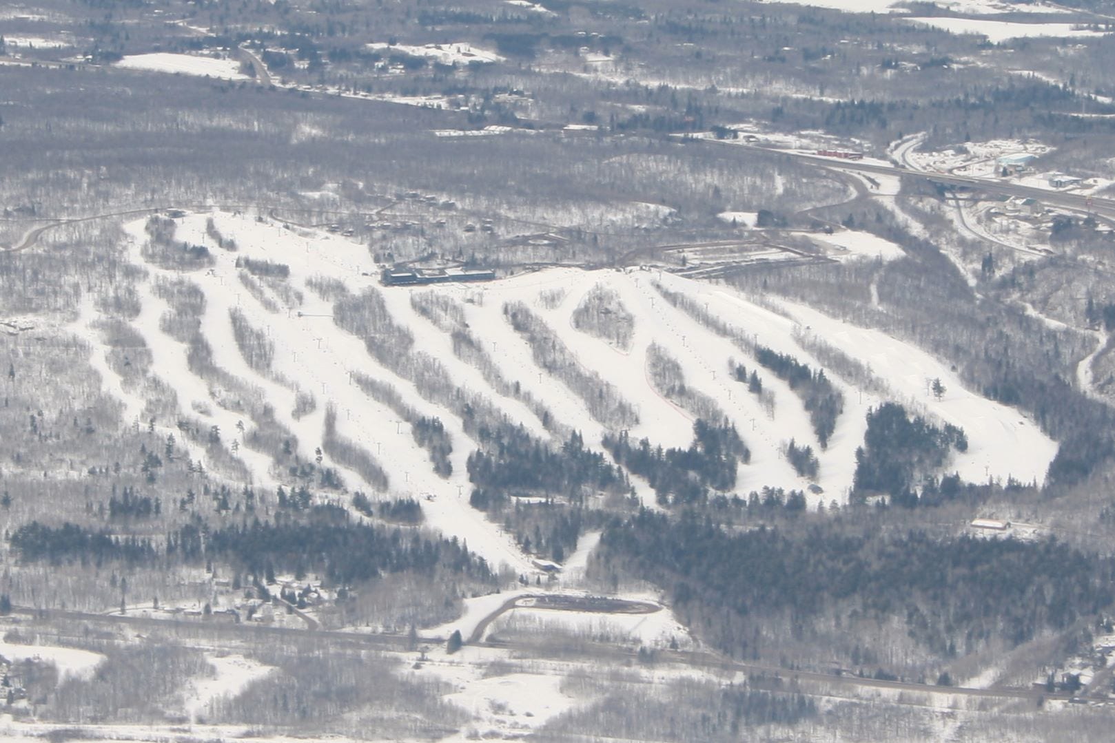 Aerial view of a snow-covered ski hill with many trails, lifts and a lodge above a wooded valley