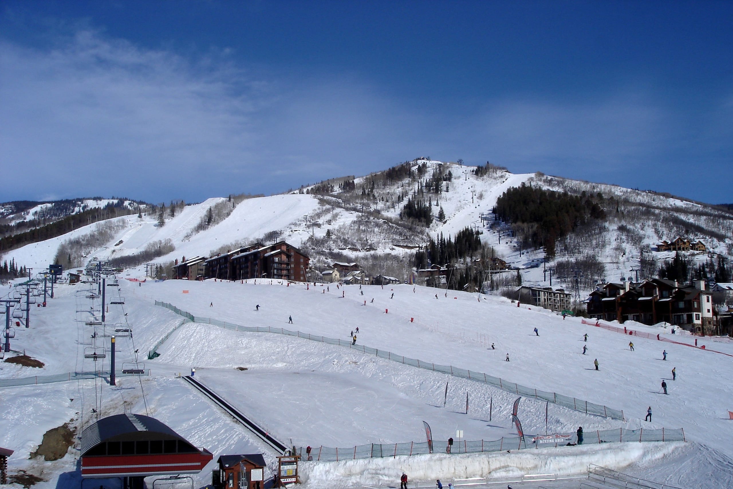 Snow-covered beginner slopes, chairlift and base village buildings below a forested ski mountain at Steamboat