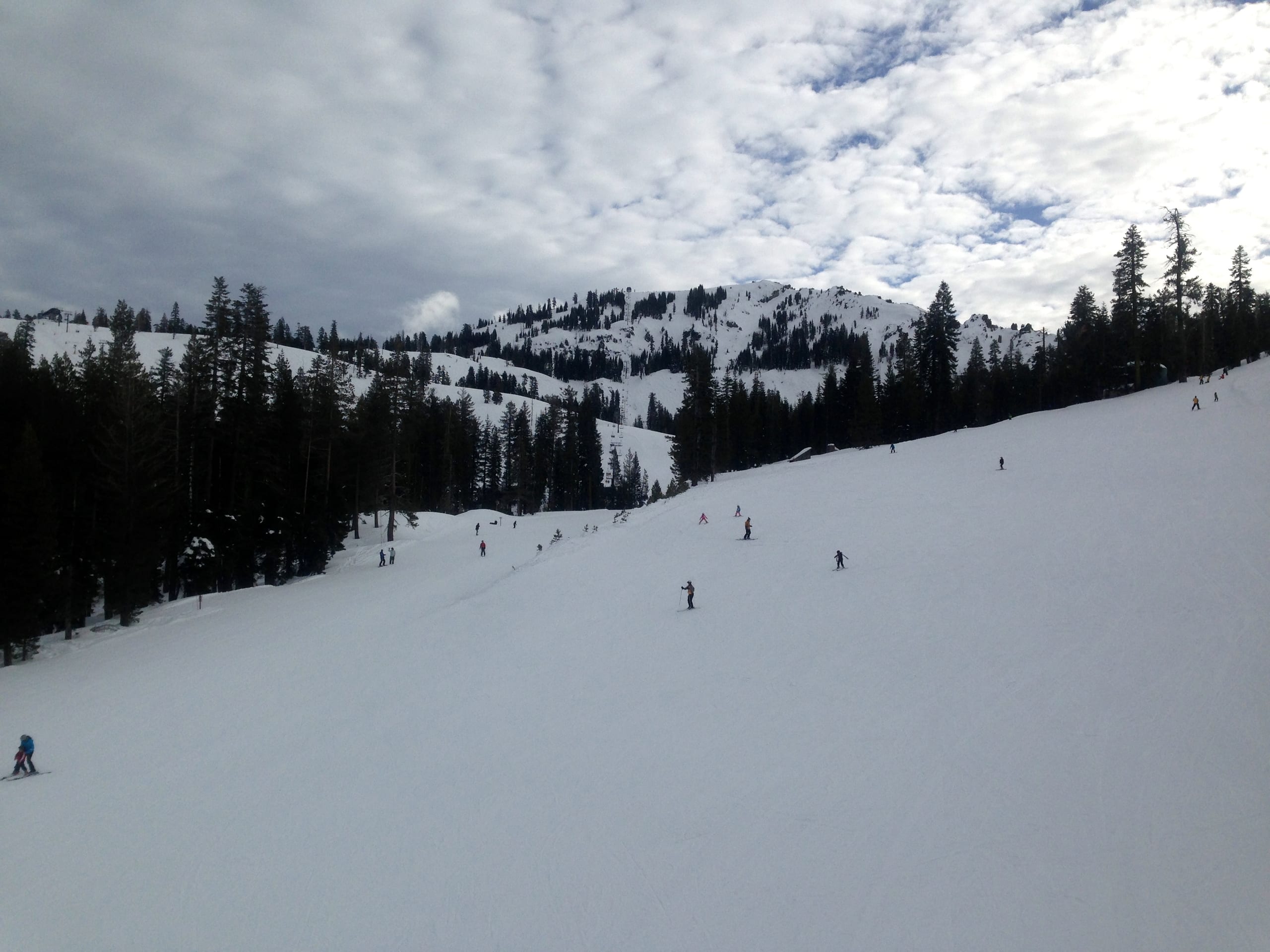 Skiers descend a wide snow-covered run lined with conifers, with a broad snowy ridge beyond under cloudy sky