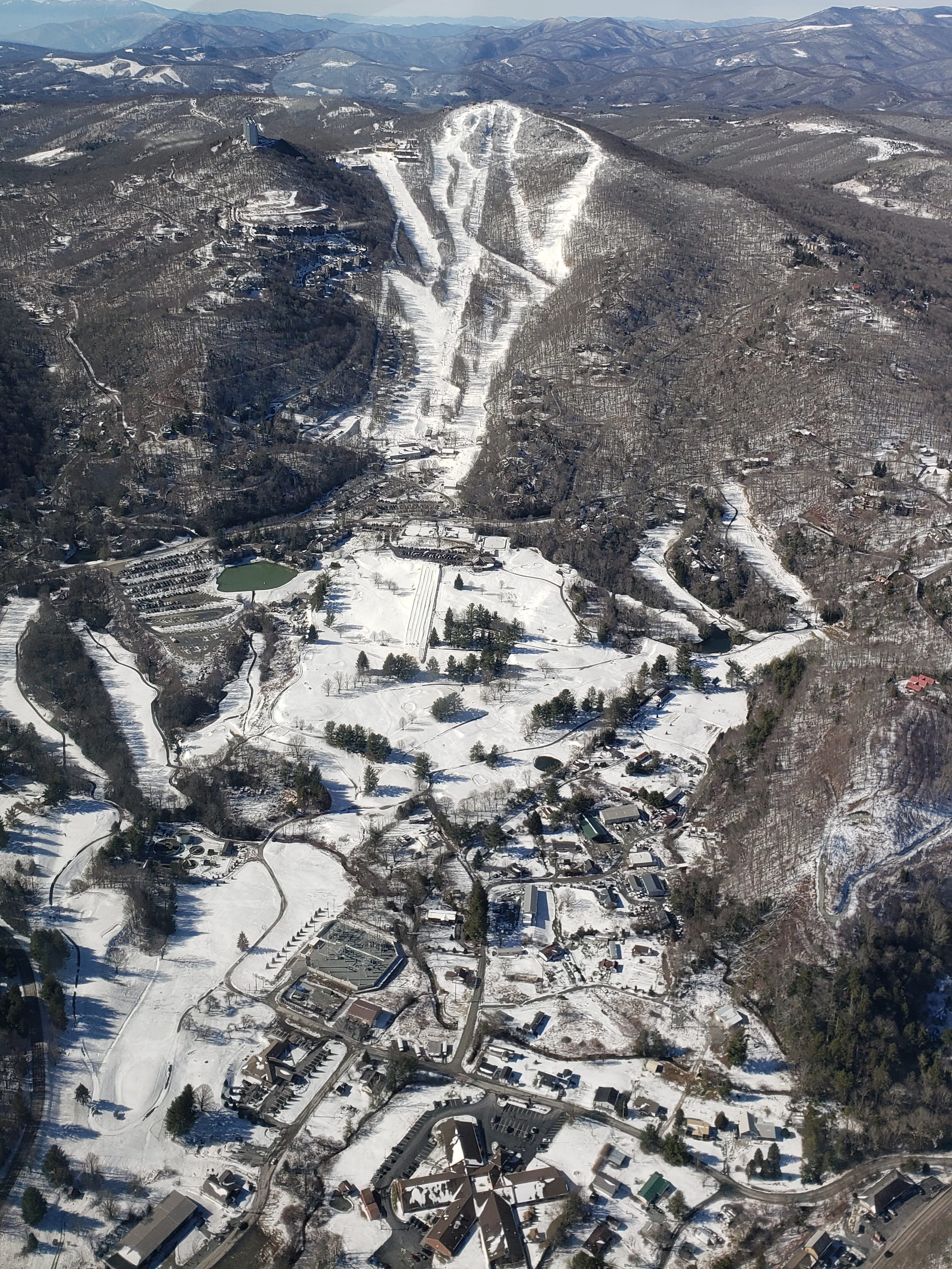 Aerial view of snow-covered ski runs cut down a wooded ridge above a snowy village and golf course.