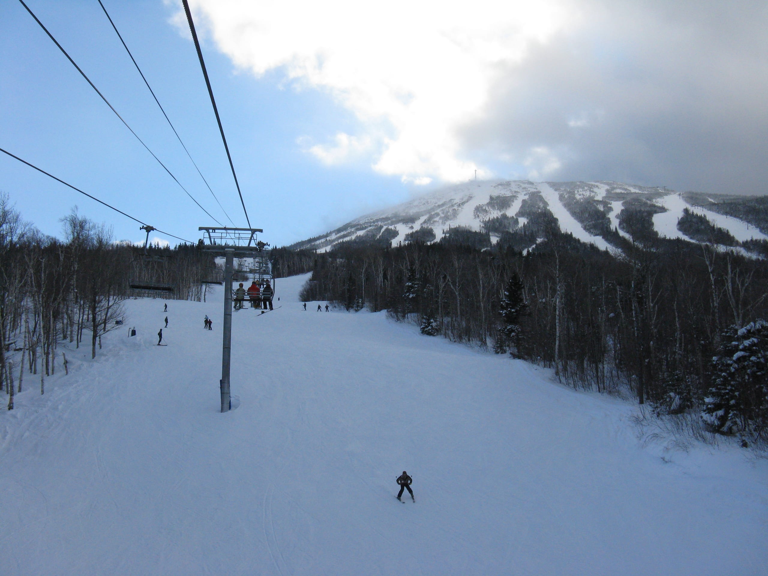 View from a chairlift over a wide snowy trail with skiers, Sugarloaf's cloud-capped summit and trails beyond