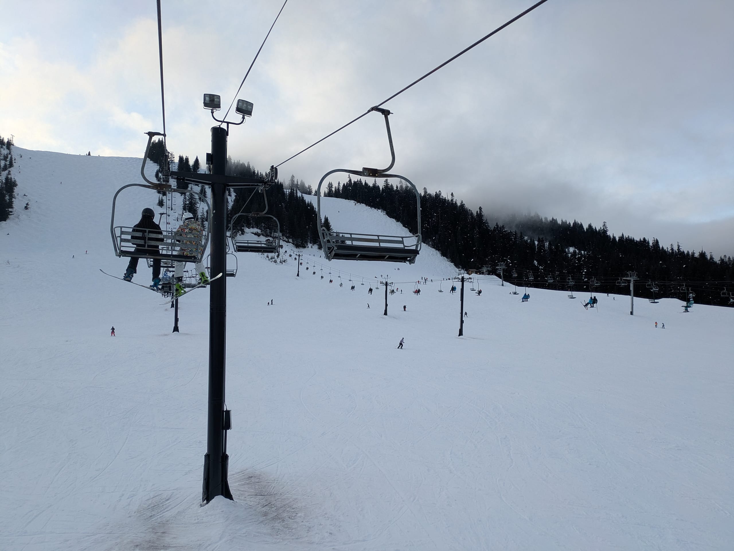 Chairlift towers over a broad snow-covered slope with skiers and forested ridge under overcast sky