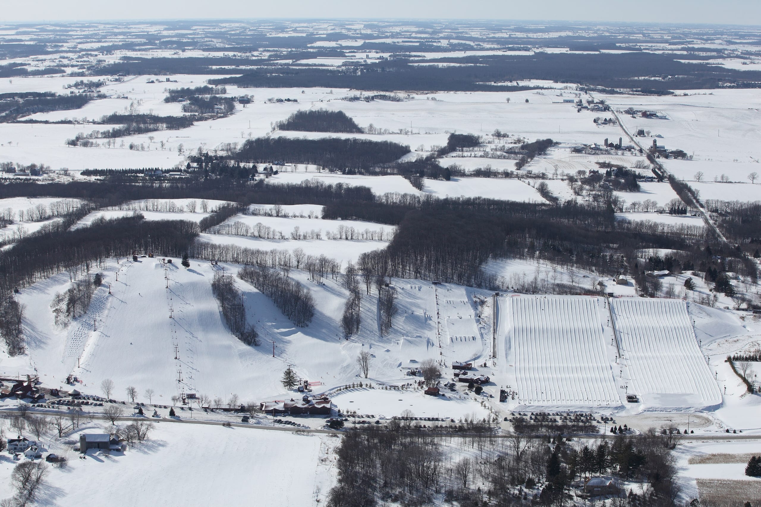 Aerial view of a small snow-covered ski hill with runs, lifts and tubing lanes amid Wisconsin farmland
