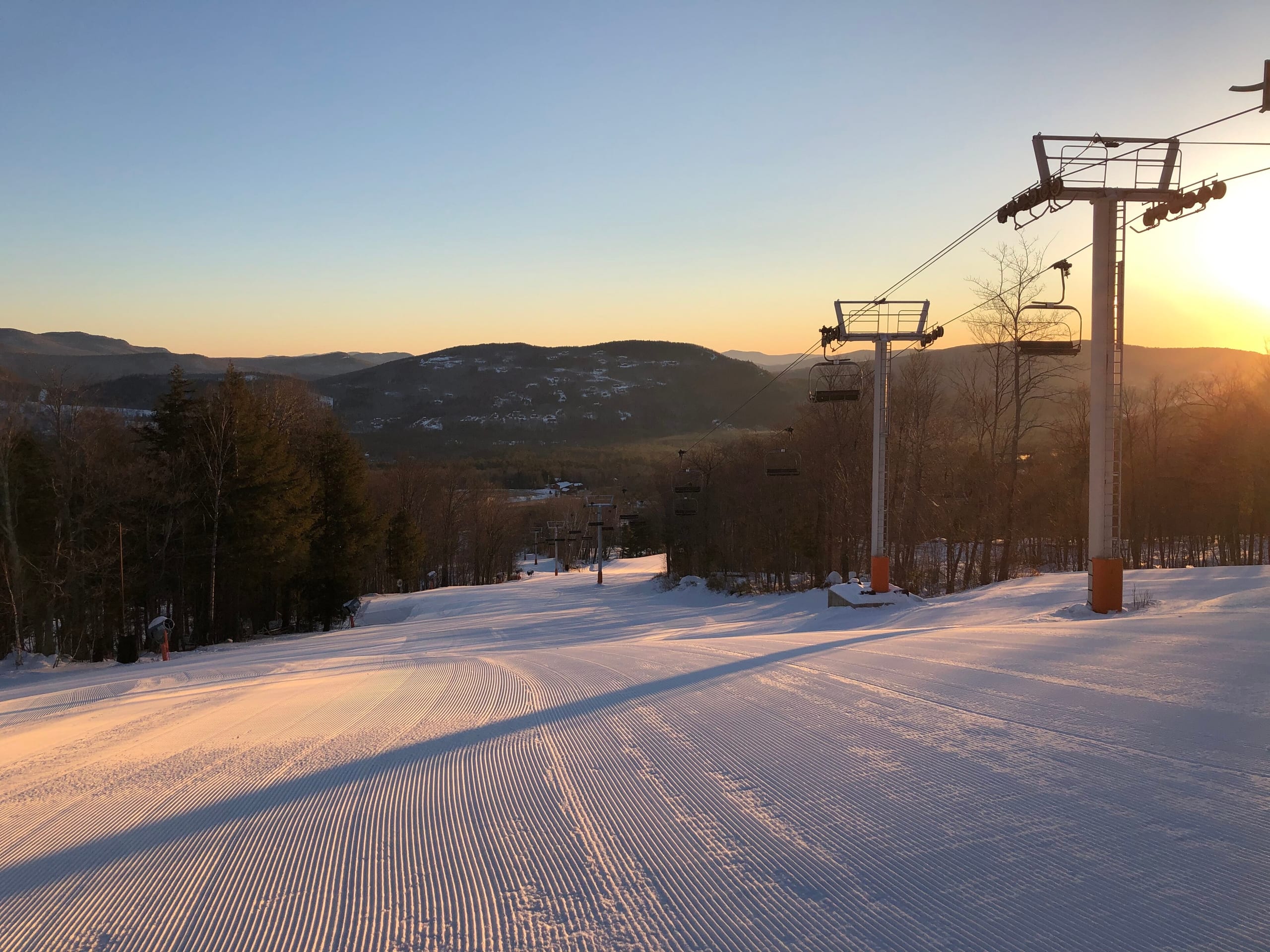 Freshly groomed ski run with a chairlift at sunrise, forested hills beyond