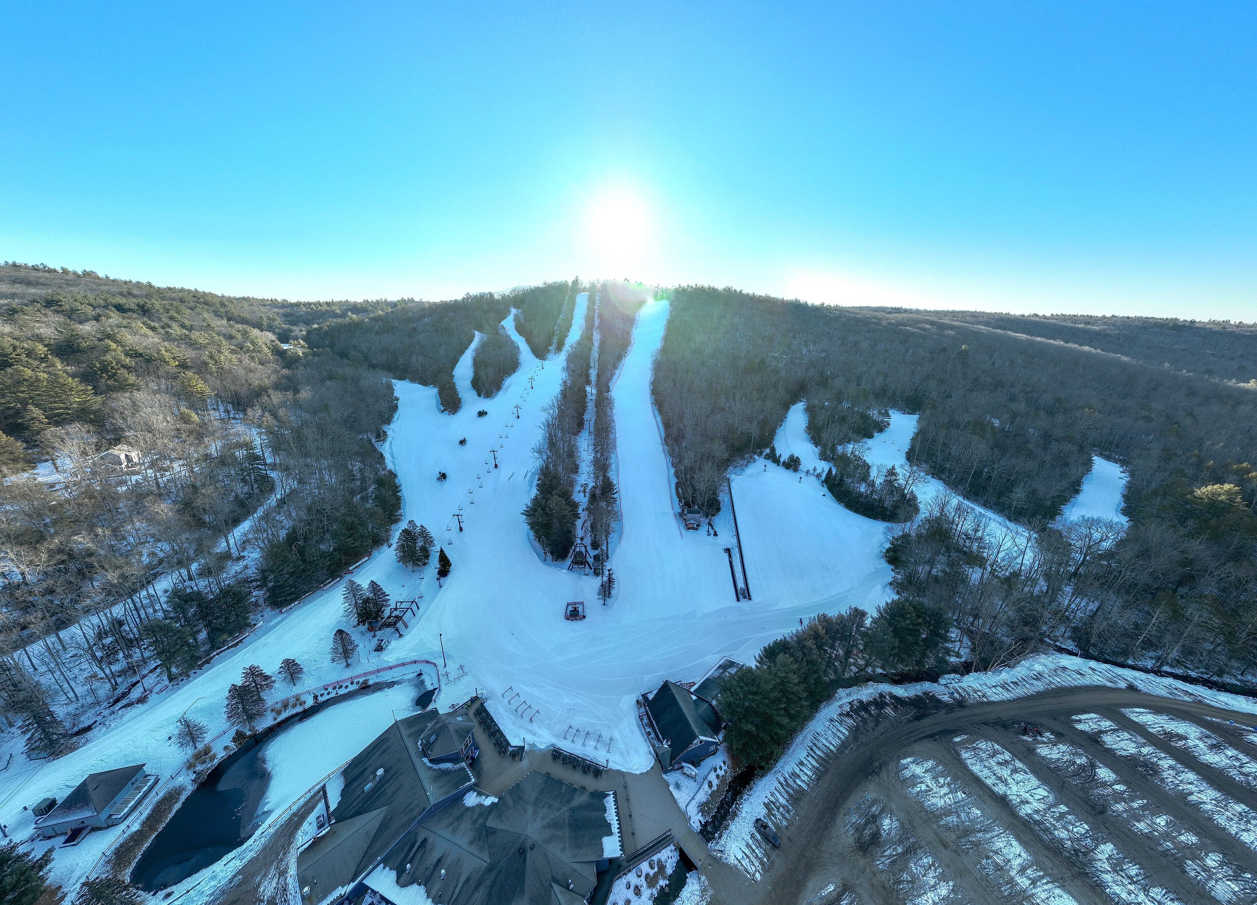 Aerial view of a small ski area with snow-covered trails, base lodge and parking lot below wooded hills