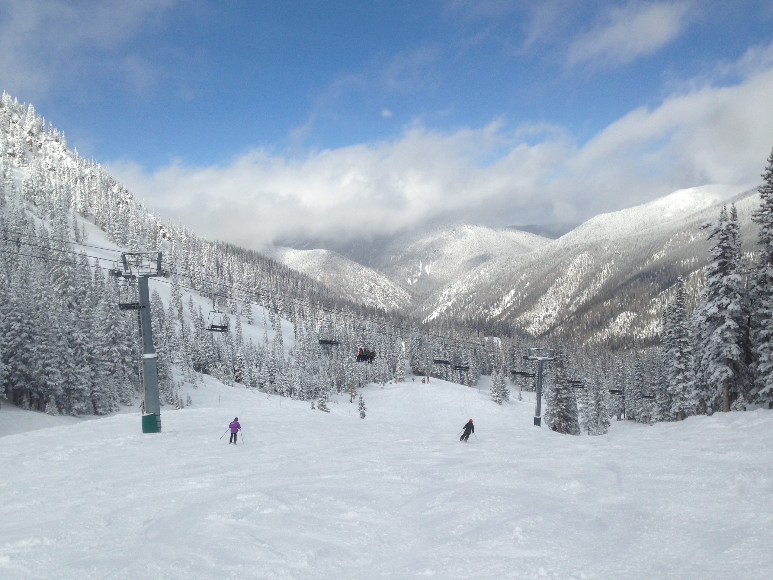 Skiers descend a snow-covered run beside a chairlift with forested peaks beyond at Taos Ski Valley