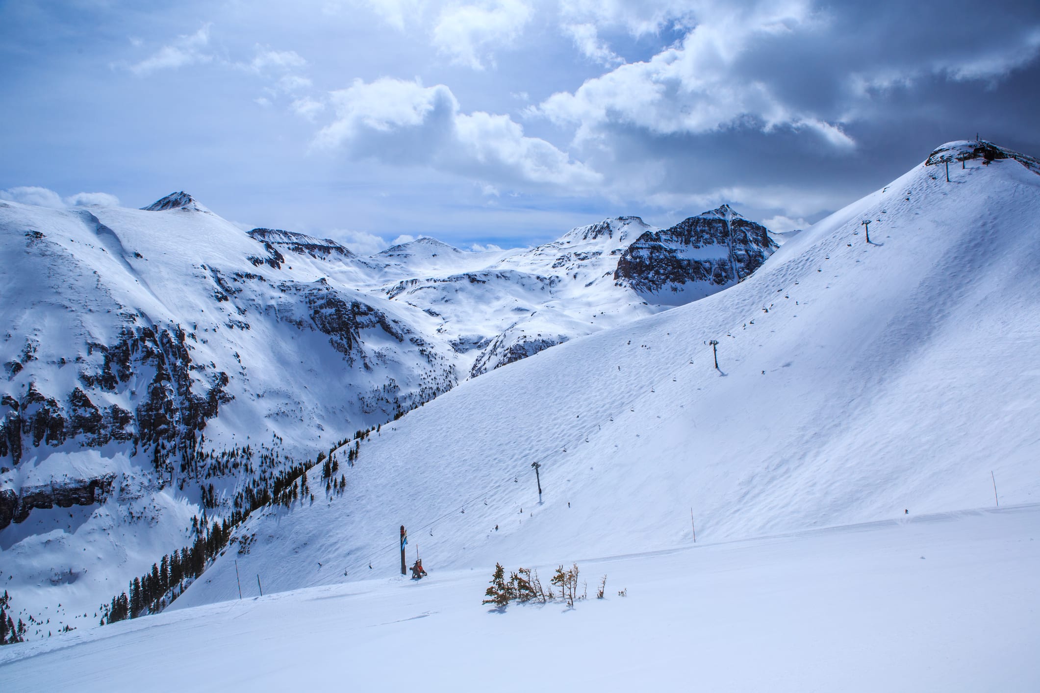 Snow-covered ski slope with a chairlift line climbing a ridge, surrounded by high peaks under cloudy sky