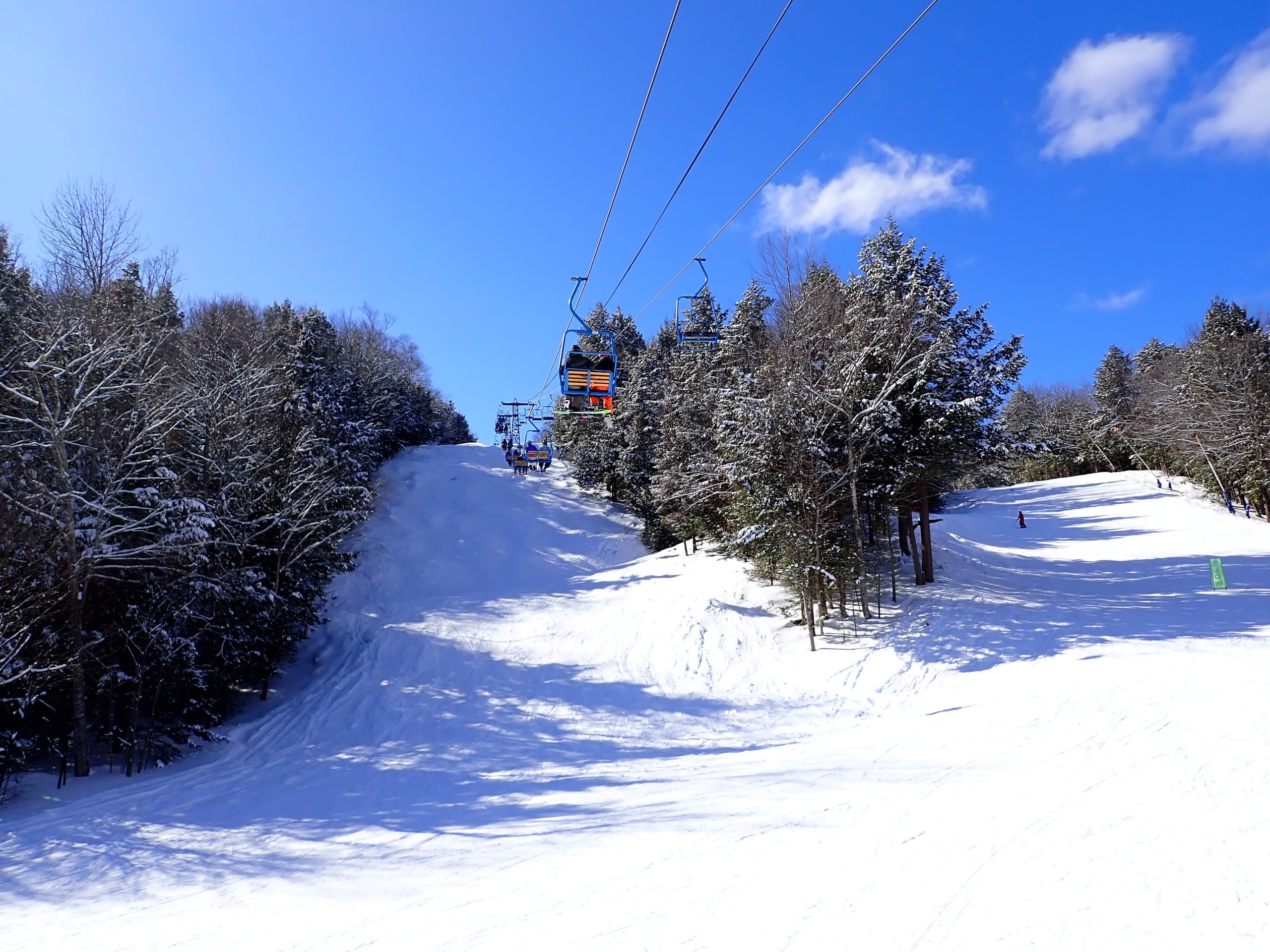 Snow-covered ski trails lined with evergreens beneath a double chairlift under a blue sky