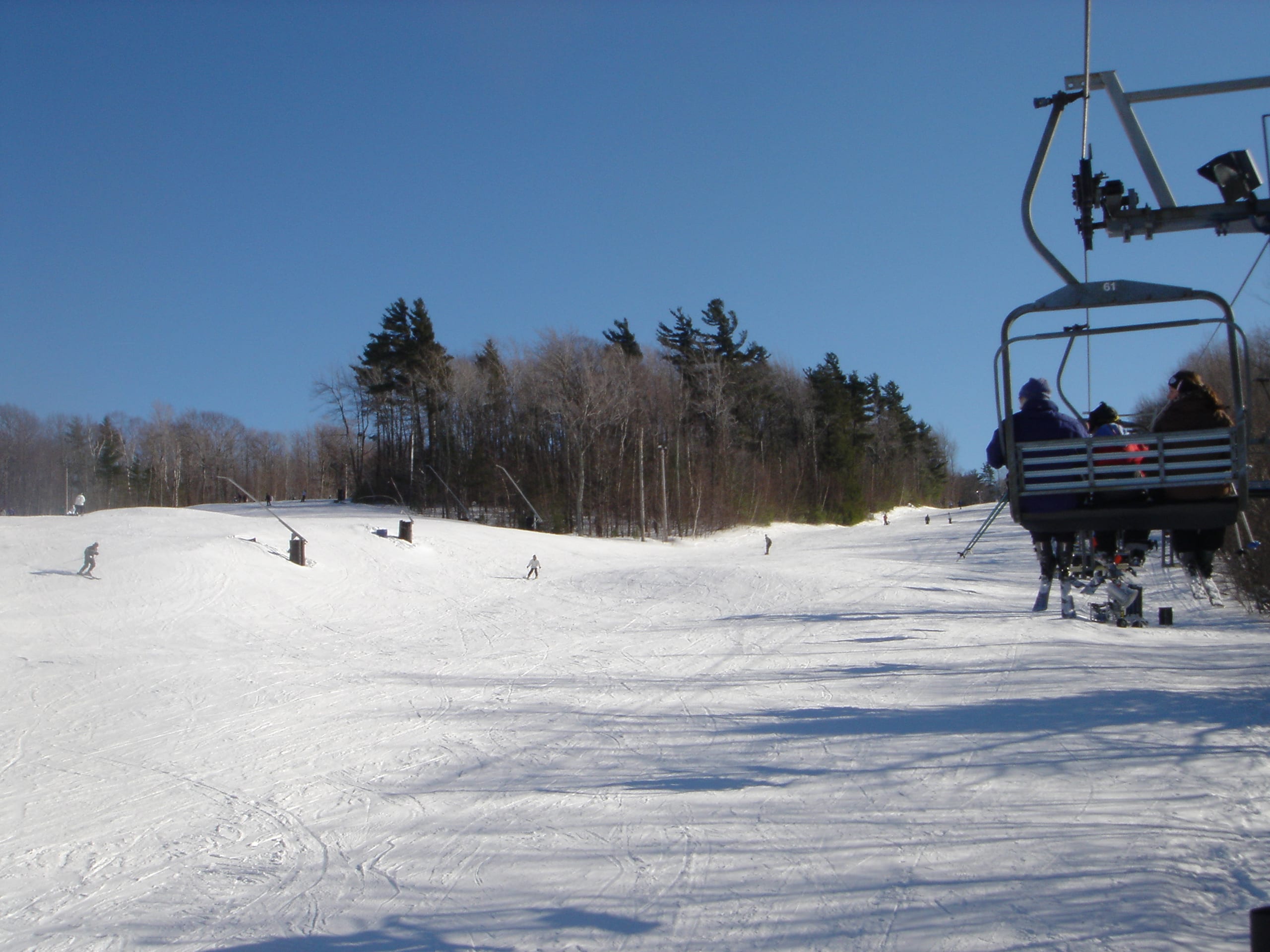 Skiers on a wide snowy slope beside a chairlift carrying riders under a clear blue sky