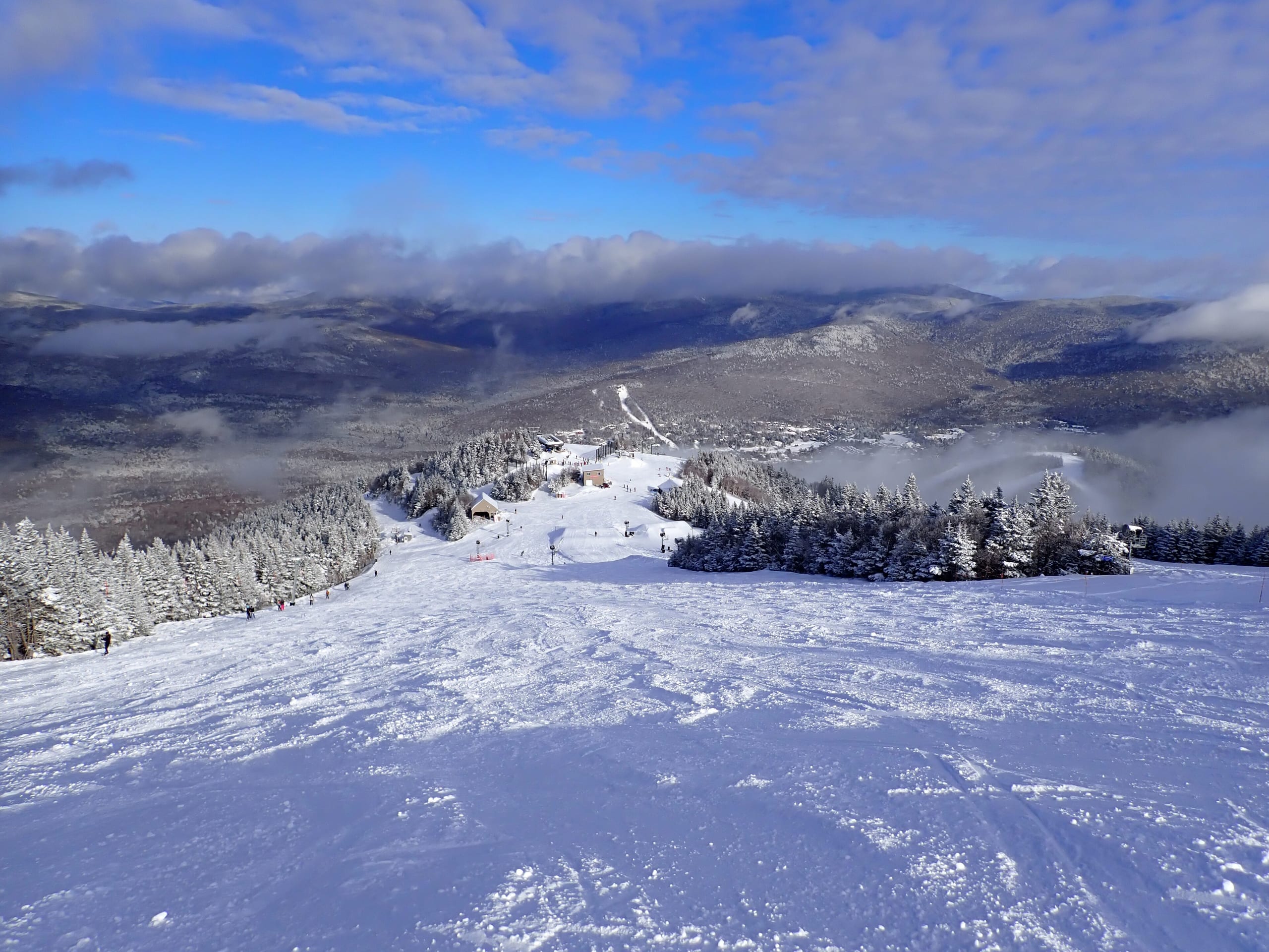 Snow-covered run descending from the upper mountain toward a mid-mountain lodge, with forested peaks beyond