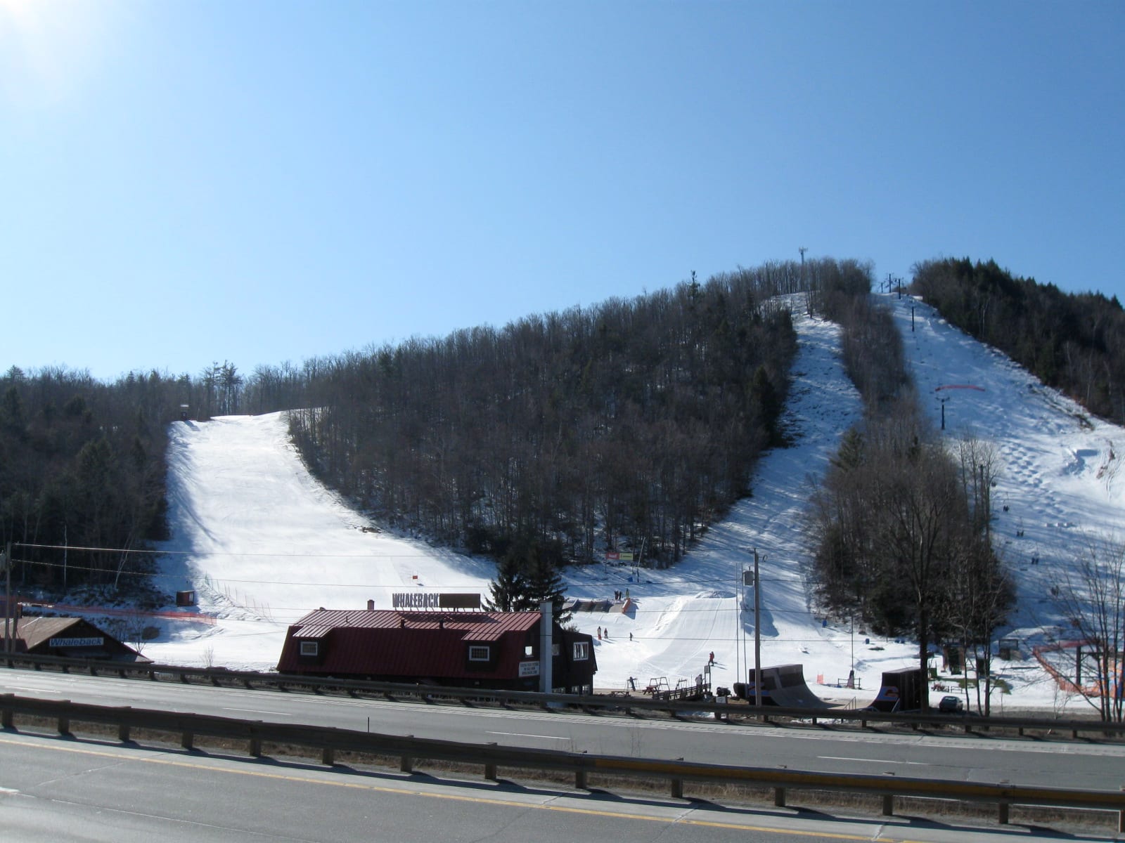 Snow-covered ski trails and a red base lodge at Whaleback Mountain, seen from across a highway