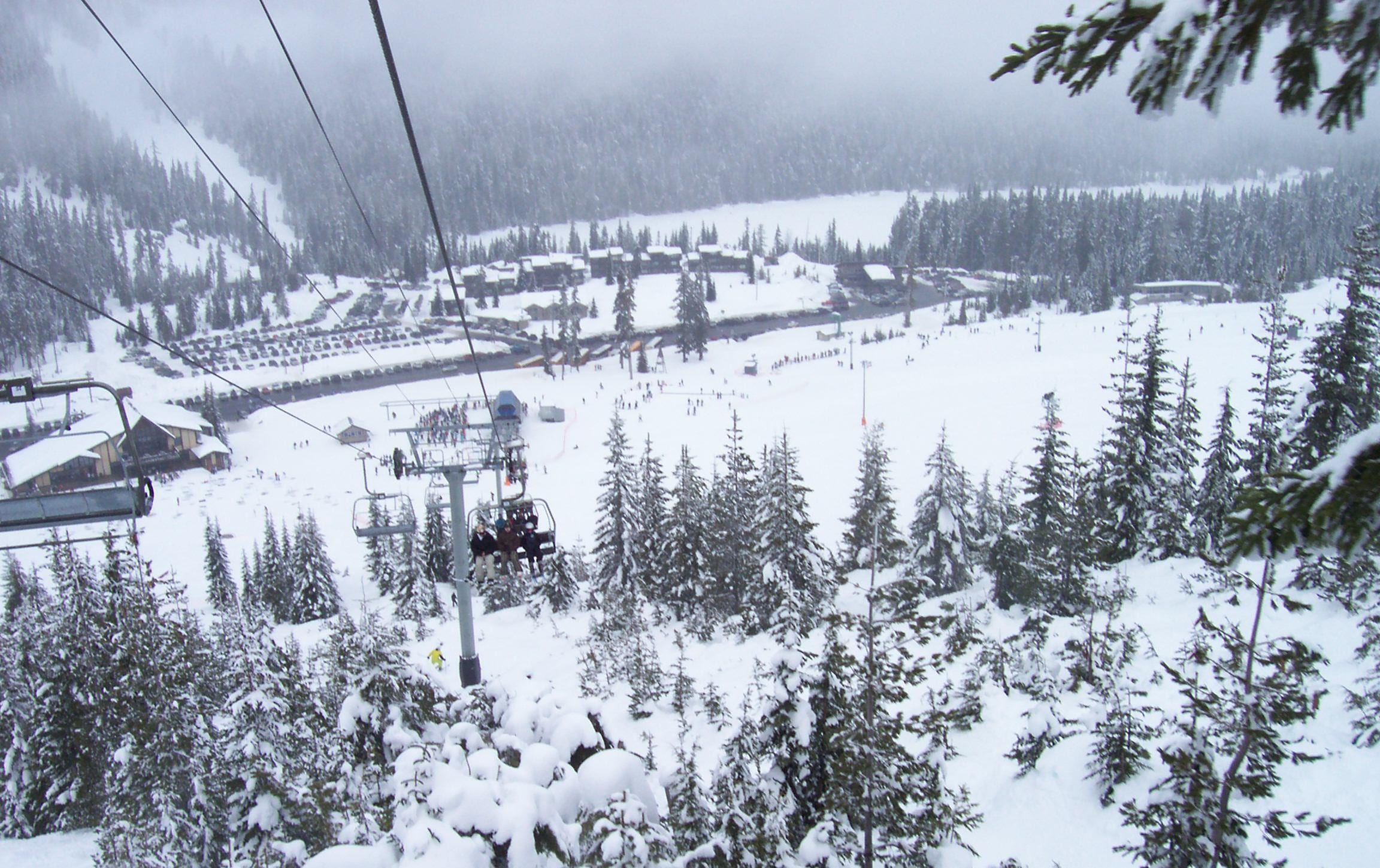 View down a snowy chairlift line toward the White Pass base area, lodge and parking lot in flat light