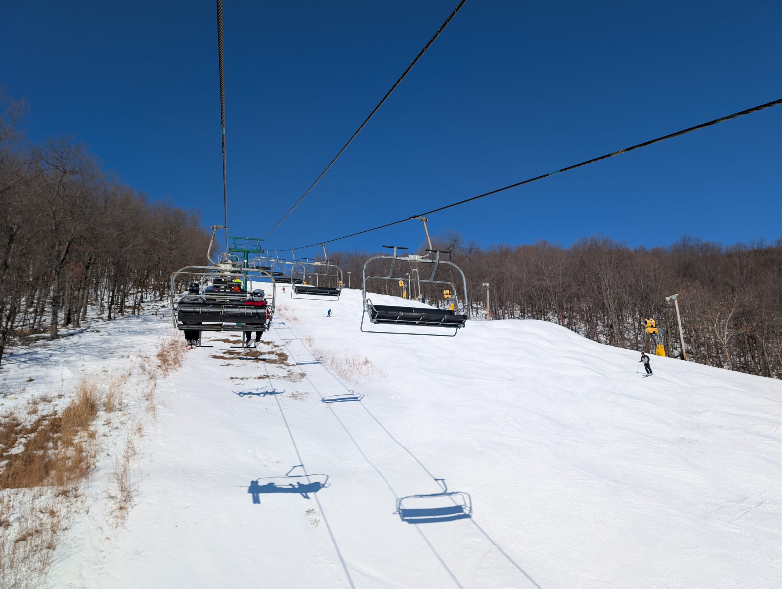 View from a chairlift up a snow-covered ski slope with bare winter trees and skiers under blue sky