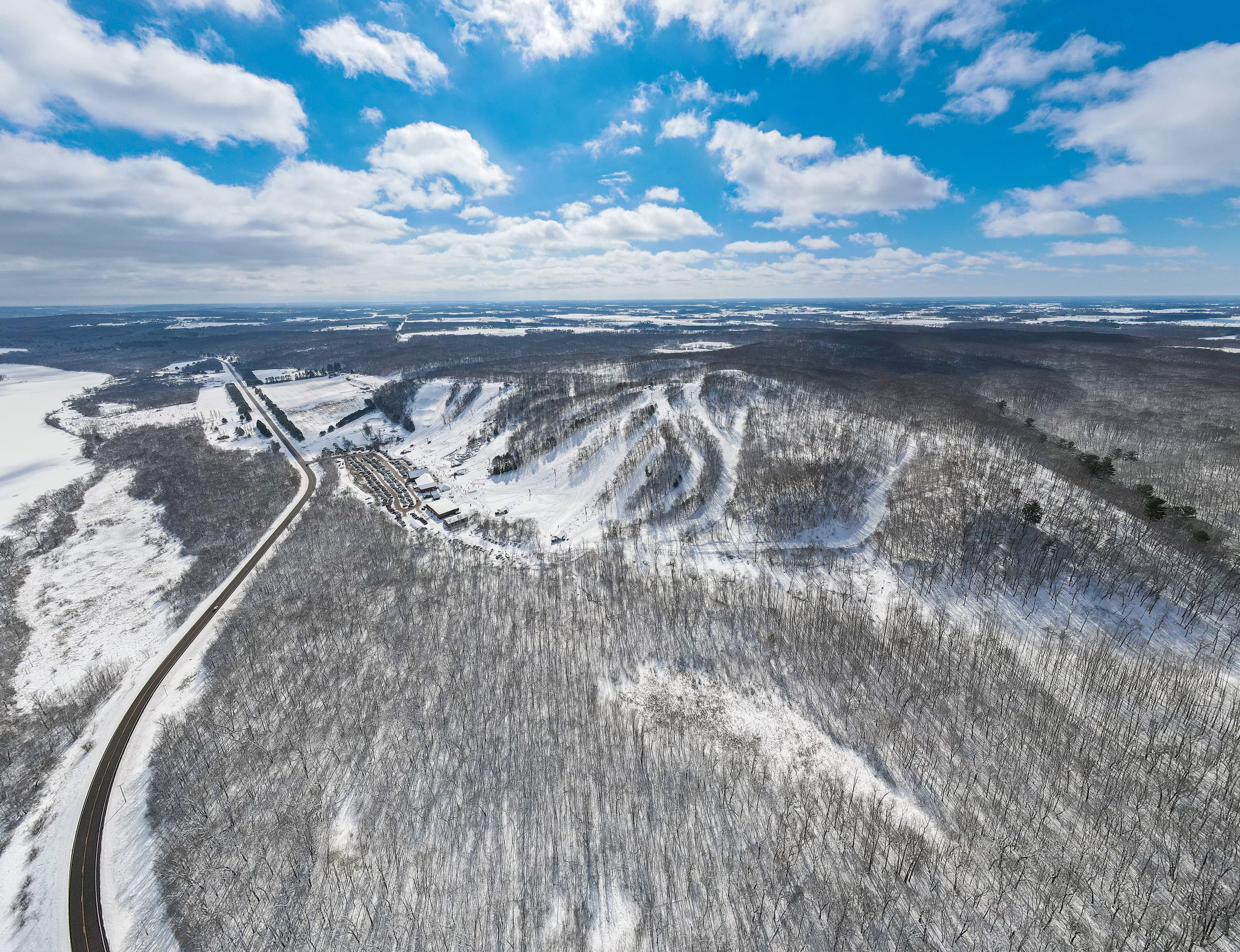 Aerial view of a small snow-covered ski hill with trails, base lodge and parking beside a frozen river valley
