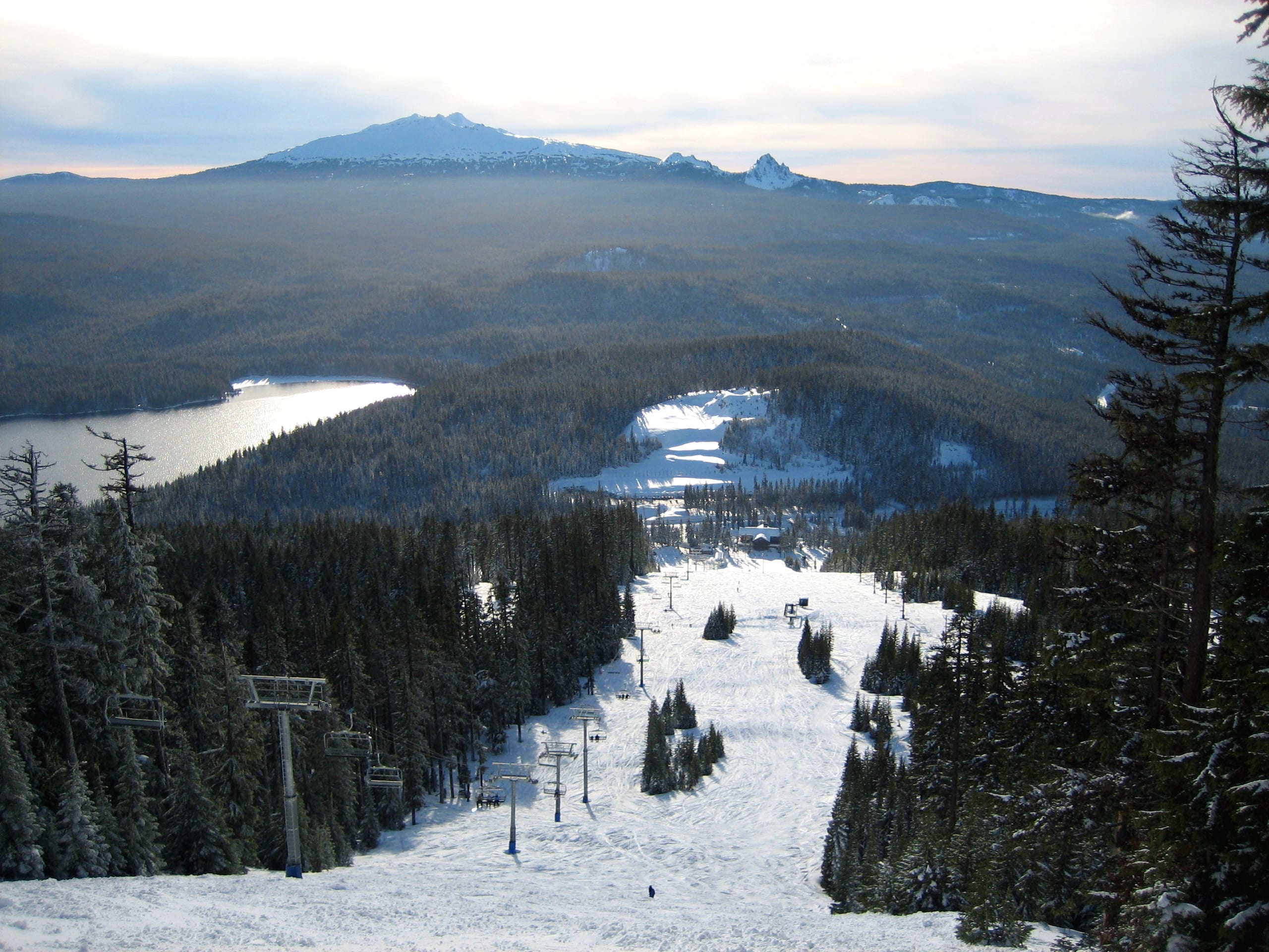 View down a snowy ski run with chairlifts toward a base area, a lake and forested peaks beyond