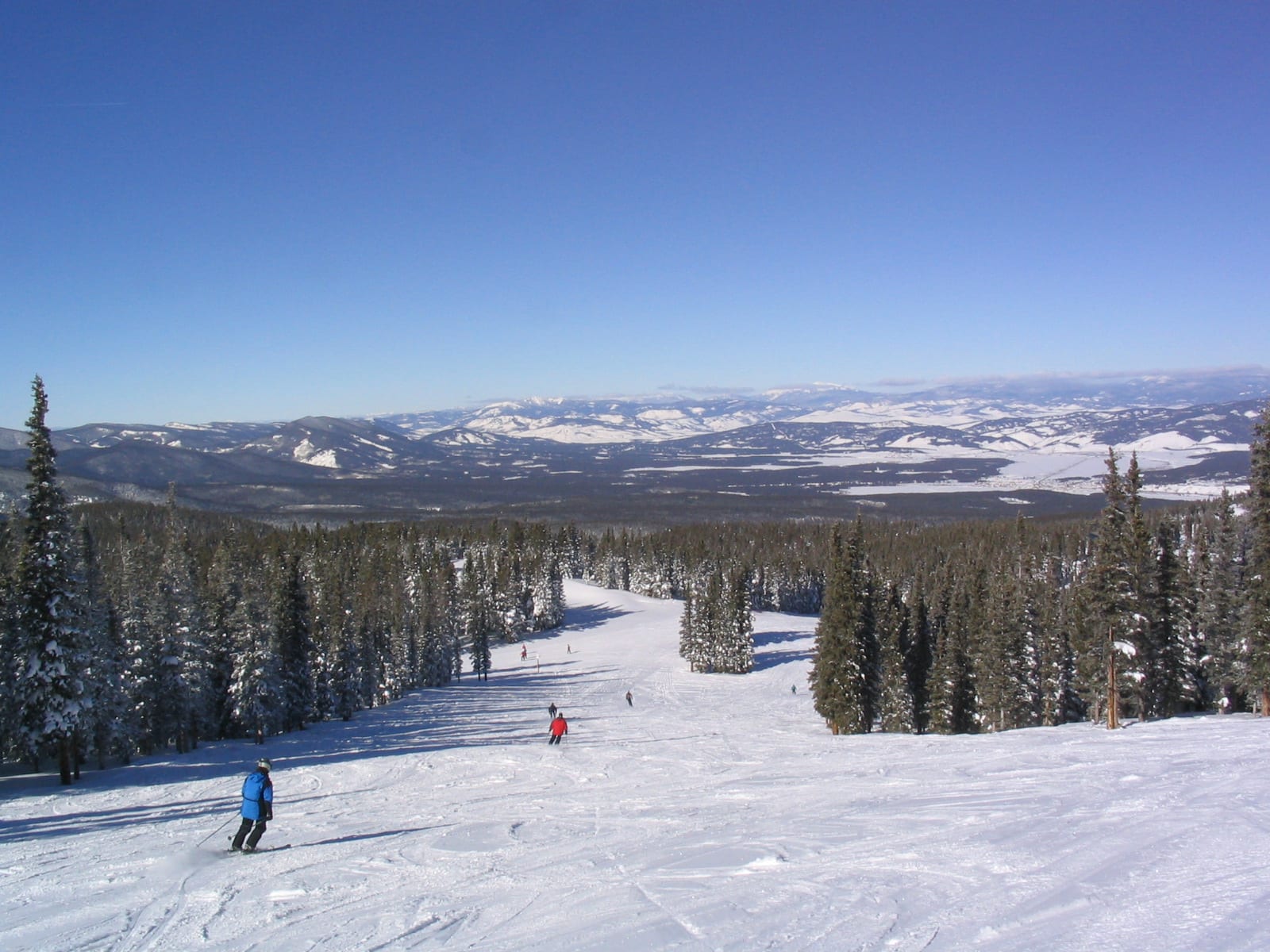 Skiers descending a wide groomed trail lined with snowy conifers, with a broad snow-covered valley beyond