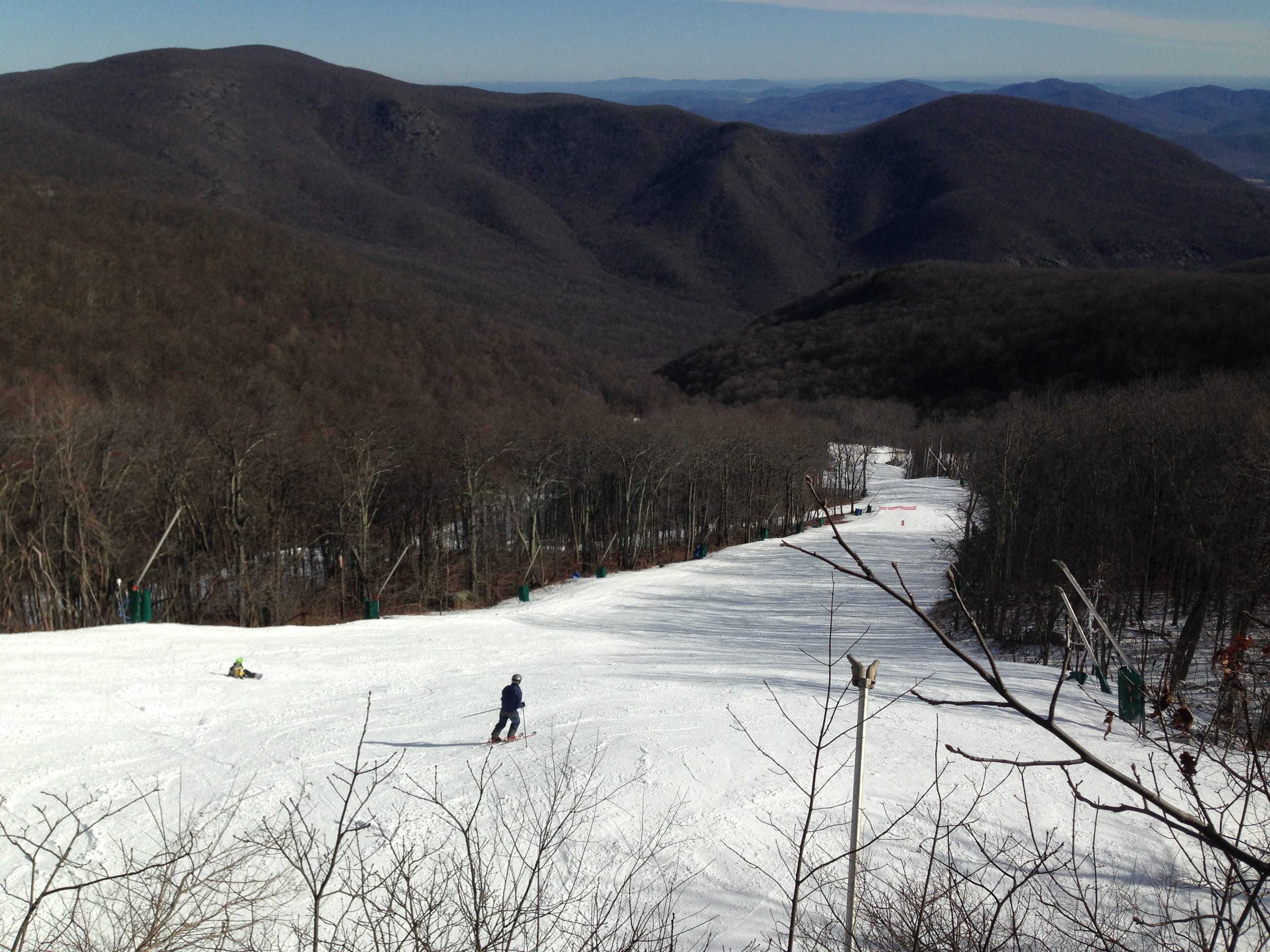 Skiers descend a groomed run lined with bare trees, with forested Blue Ridge ridges beyond