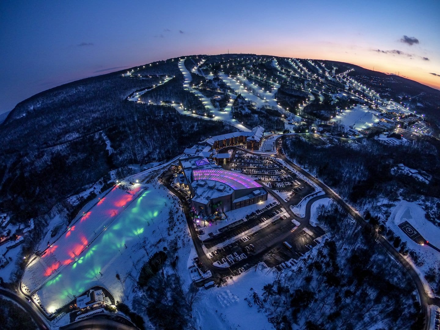 Aerial dusk view of a snowy ski hill with floodlit trails above a base village and glowing tubing park.