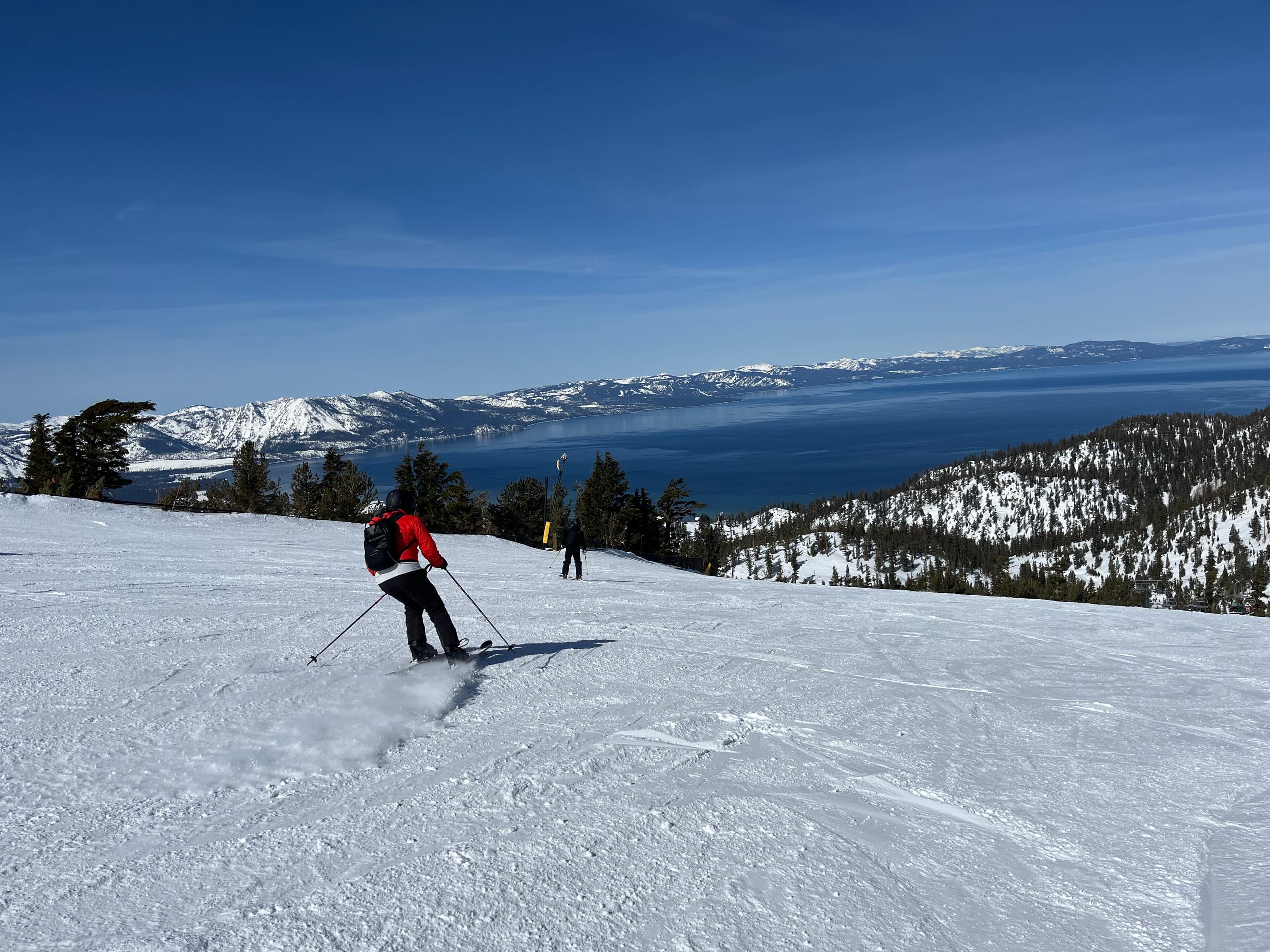 A skier in red descends a groomed snow run high above Lake Tahoe under a clear blue sky at Heavenly.