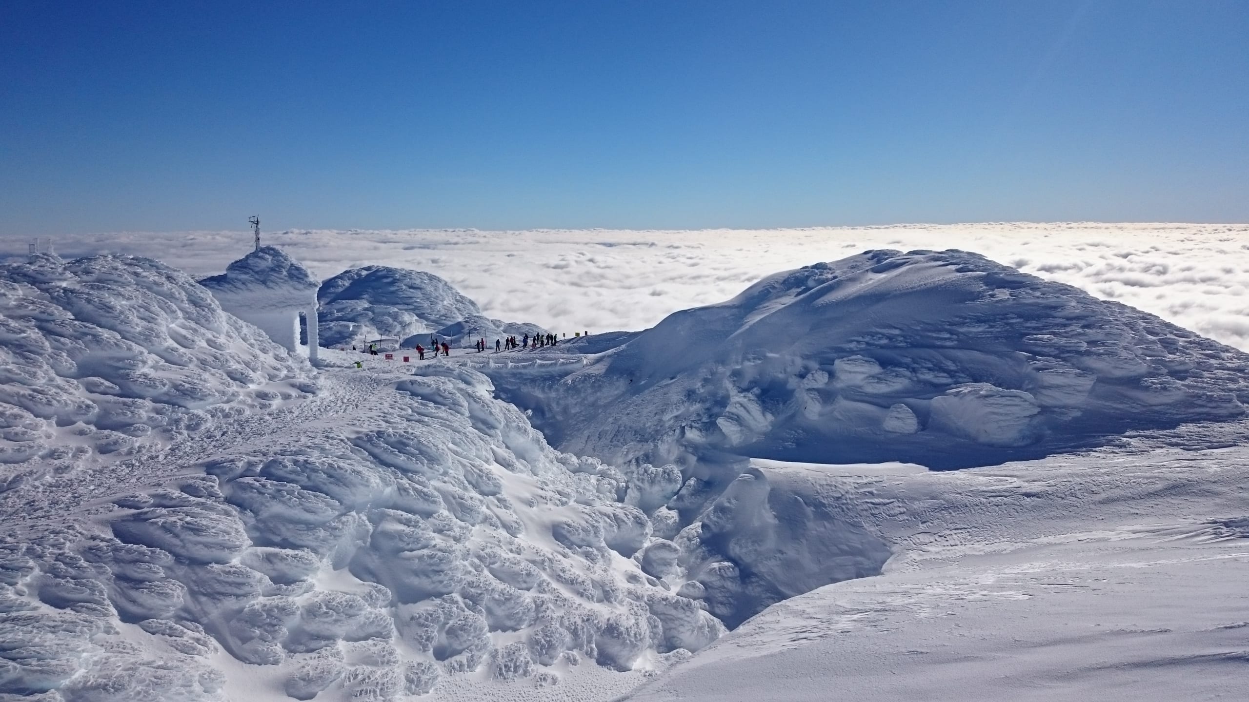Rime-covered summit ridge with a small hut and a line of tiny figures above a sea of clouds.