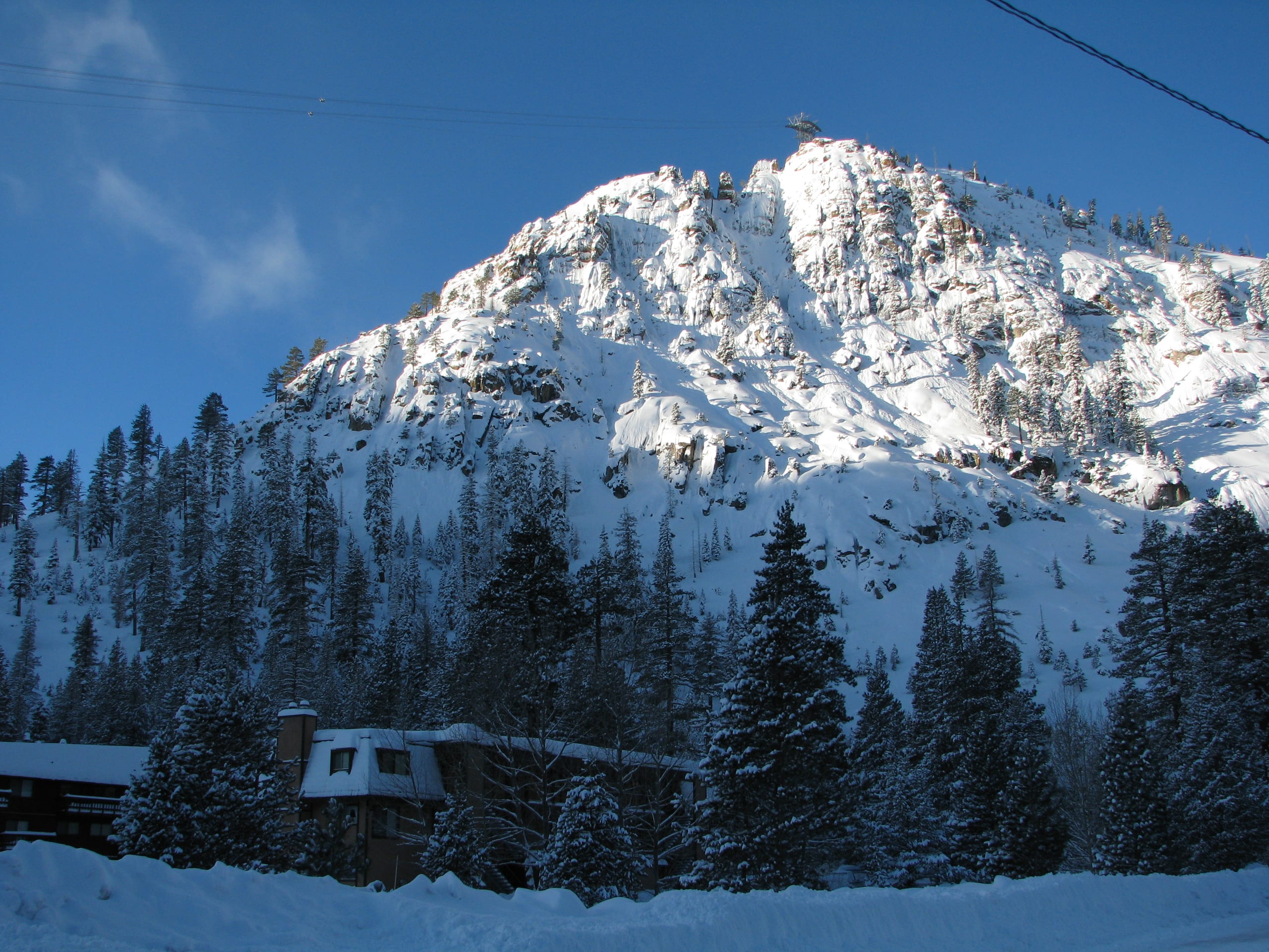 Snow-covered rocky peak with a cable car station on top, above pines and a building at Squaw Valley