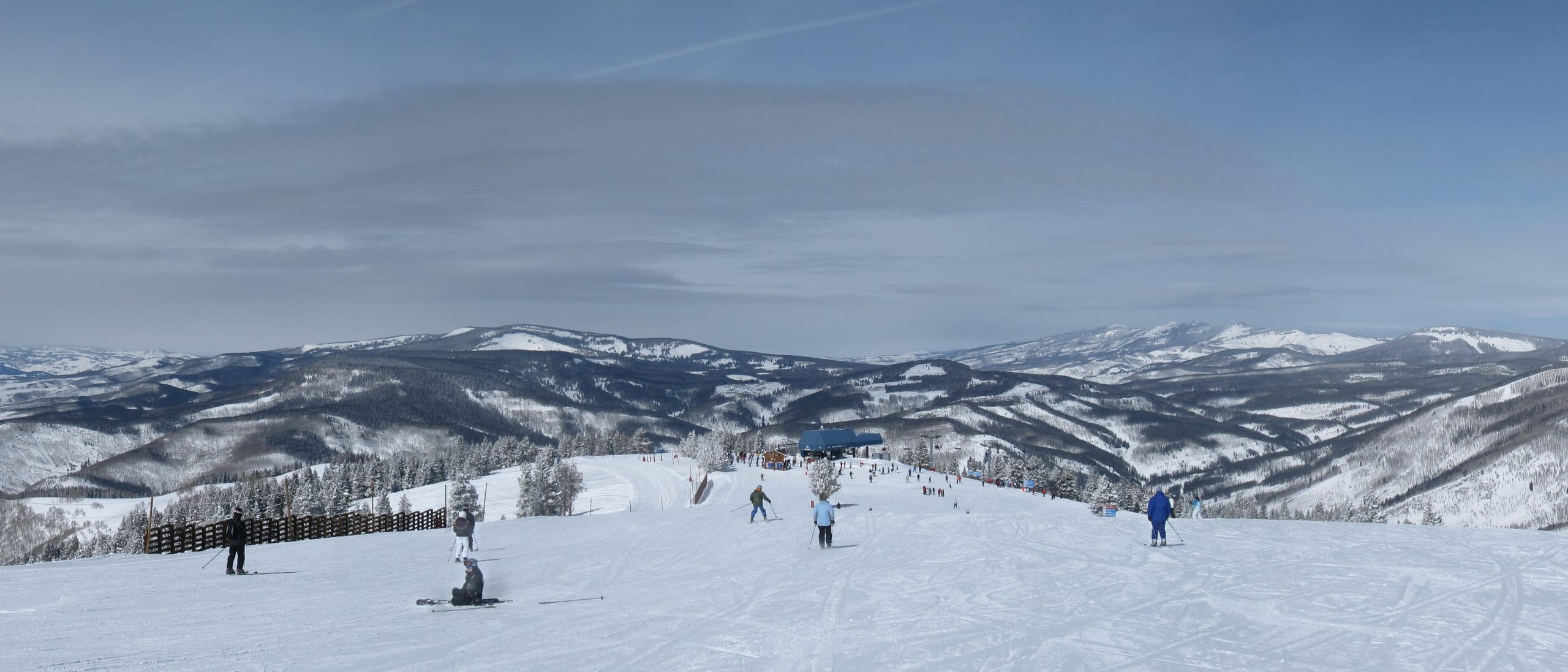 Skiers on a wide groomed run atop a ridge, with snow-covered forested mountains stretching to the horizon.